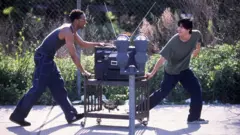 Dois homens empurrando uma televisão em um carrinho