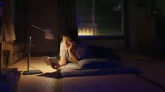 Homem lê um livro à noite, deitado no chão de casa, com uma luminária