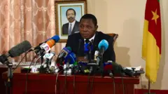 Mínísítà fọ́rọ̀ abẹ́lé ní Cameroon, Paul Atanga Nji wọ kóòtù dúdú pẹ̀lú ṣáàtì funfun lábẹ́ àti Táì aláwọ̀ búlúù ní ọrùn rẹ̀ . Ọ̀pọ̀lọpọ̀ gbohùngbohùn wà níwájú rẹ̀, tí àwòrán ààrẹ Cameroon, Paul Biya wà lẹ́yìn rẹ̀.