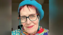 A selfie of Mary Thornton. She has short red hair and blue eyes. She is wearing a bright blue beret and a colourful shawl. She also wears glasses with a blue and orange frame. She is slightly smiling.