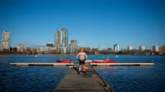 James está em uma rampa na represa. Ele está usando um gorro laranja e shorts de banho, além de meias de banho. O céu está azul e há prédios altos ao longe, além da beira da água.