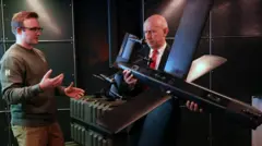 Министр обороны Великобритании Джон Хили держит в руках большой черный беспилотник, о котором ему рассказывает мужчина в военной форме