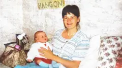 Linda sorri sentada em um sofá enquanto segura sua filha bebê, Cat. Ela tem cabelos escuros e veste um cardigã branco e azul. Cat usa um suéter branco, shorts vermelhos e meias azuis compridas.