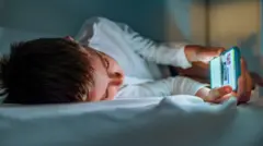 Menino deitado na cama, olhando a tela do telefone celular
