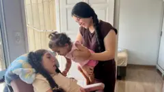 Inka segura Ester no colo enquanto ela toca o rosto da irmã Sophia, que está em uma cadeira de rodas