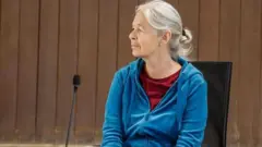 Женщина с седыми волосами сидит перед микрофоном в синей толстовке на молнии и красной майке