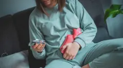 A woman is sitting comfortably on her couch and with a hot water bottle pressed against her stomach and a packet of pills in her hand.