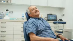 Juan Ramón Soto é um homem idoso, com cabelos grisalhos. Ele está sentado na cadeira de um consultório odontológico e usa uma camisa azul