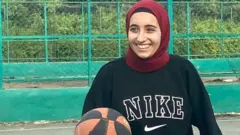 Una mujer con velo sonríe mientras mantiene un balón de baloncesto en la mano.
