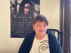 Andrea está sentada e olha para a câmera. Em uma parede clara ao fundo, um pôster do filme "A Mulher na Fila" está colado, mostrando uma mulher na fila com a sombra de uma cerca projetada em seu rosto.