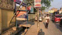 একটি রাস্তায় গাড় চলছে এবং একজন তরুণ পথচারী হেঁটে যাচ্ছেন। রাস্তার পাশে হিন্দিতে লেখা নাথনগর থানা