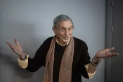 Haldun Dormen, siyah kazak ve kahverengi, desenli bir atkı giymiş halde ellerini iki yana a&ccedil;ıp, g&uuml;l&uuml;mserken 