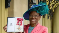 Prof. Dame Ijeoma Uchegbu berdiri di latar depan, mengenakan jaket koral, blus biru muda, dan topi biru muda bertepi samping. Ia tersenyum. Ia memegang sebuah kotak panjang terbuka berisi lencana Dame Commander of the Order of the British Empire. Hiasannya berupa pita koral lebar bertepi perak dan dihiasi salib panjang berwarna emas, perak, dan biru muda.