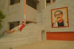Um mural exibe a imagem de Sheikh Mujibur Rahman em um muro. Em uma parede ao lado, uma menina usando protetores auriculares está deitada