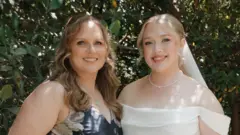 Hannah and her mother at Hannah's wedding. Her mother is wearing a blue dress with white flowers. Hannah on the right is dressed in a white wedding dress and veil