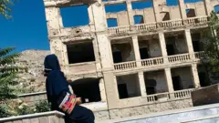 Vue latérale de Nadia portant un hijab noir avec des manches brodées de couleurs devant un bâtiment endommagé.