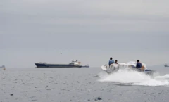 Üç kişinin içinde olduğu bir deniz motoru hızla ilerliyor. Arkada büyük bir tanker var.