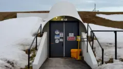 Two black doors with warning signs built into a snow covered hill, with some dead grass showing through. They are covered by a white arc-shaped cover and a path, with white concrete walls, is paved in front of them