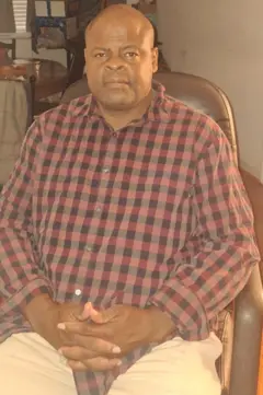 Ron Bishop sentado em uma camisa xadrez, mãos entrelaçadas, olhando para a câmera