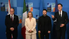 Premier Włoch Giorgia Meloni stoi w centrum zdjęcia w beżowym garniturze, obok niej stoją premier Wielkiej Brytanii i przywódca Ukrainy, a po prawej Mark Rutte