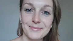 A imagem mostra uma mulher em um retrato em close, olhando diretamente para a câmera e sorrindo de forma discreta. Ela tem olhos azuis, cabelos castanhos claros penteados para o lado e usa brincos de pérola.