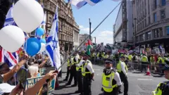Polis memurları, bir sokakta iki protestocu grup arasında duruyor. Solda, İsrail bayrakları, bir Birlik Bayrağı (Union Jack) ve beyaz ile mavi balonlar görülüyor. Sağda ise metal bir bariyerin arkasında ayrı bir kalabalık, Filistin bayrakları ve çeşitli protesto pankartları tutuyor. Arka planda tarihi binalar ve parlak mavi bir gökyüzü var.
