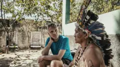 Dom et la chamane sont assis dans la partie extérieure de la maison ; elle parle tandis qu'il écoute.