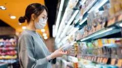 Uma jovem está na seção refrigerada de um supermercado, segurando um pote de iogurte orgânico fresco e saudável, lendo o rótulo nutricional e verificando os ingredientes no verso