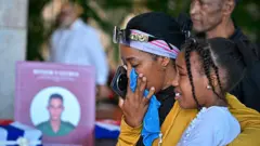 Uma mulher enxuga as lágrimas enquanto abraça uma menina, com a foto de um soldado ao fundo.