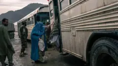 تصویر یک زن مهاجر افغان که چادری آبی به سر دارد. او درحال سوار شدن به بسی حامل مهاجران است. در تصویر دو مرد مهاجر دیگر نیز دیده می‌شوند یکی شان سوار بس شده تنها لباس هویدا است و دیگری در عقب زن ایستاده. یک سرباز نیز در تصویر دیده‌شود. هوا سرد است و زمین گل‌آلود. 