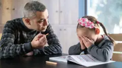 رجل يجلس إلى طاولة ممسكًا قلمًا وينظر إلى طفلة صغيرة تغطي وجهها بيديها، بينما يجلسان أمام كتاب مفتوح، في مشهد يوحي بلحظة توتر أو انفعال أثناء حلّ الواجب المدرسي.