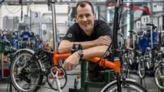 Will Butler-Adams, presidente executivo da Brompton Bicycle, com uma camisa polo preta apoiado em uma bicicleta laranja