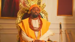 His Majesty, Ogiame Atuwatse III, Olu of Warri sitdown for im royal chair wey be red and get golden design. E wear di traditional red beaded crown wit cream color long sleeve traditional shirt and yellow flowing robe ontop. Red traditional beads dey im neck and wrist, e also carry horsetail for hand and im sword dey for di background.
