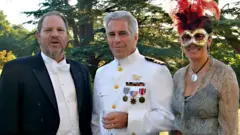 Harvey Weinstein, Jeffrey Epstein and Ghislaine Maxwell are pictured standing on the lawn at Royal Lodge on the royal estate in Windsor with trees in the background. All wear fancy dress with Weinstein and Epstein wearing suits and Maxwell is seen wearing a mask.