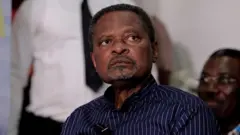 Georges Anicet Ekane, en chemise bleue foncée avec des traits verticaux, il regarde ailleurs.