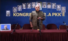 Duran Kalkan askeri kıyafetlerle lacivert bir panonun önünde. Arkasında Abdullah Öcalan dahil çeşitli kişilerin fotoğrafları asılı. 