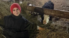 Женщина в черном пальто и красной шапочке сидит на сене рядом с коровой