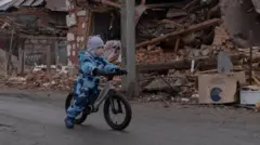 Uma criança anda de bicicleta perto de casas destruídas em Zolochiv, no oblast de Kharkiv, Ucrânia