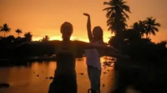 Lowri Denman e uma amiga dançam na Índia, durante o pôr do sol, em frente a um lago com palmeiras nas margens.