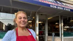 Jacqueline Tejera, vistiendo una camiseta gris y un delantal rojo, con el local de la Pizzería Montevideo detrás.