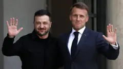 Macron y Zelensky, con trajes oscuros saludan a las cámaras, con un soldado que hace guardia a su derecha.