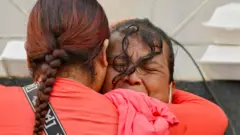 Una mujer con una camiseta manga larga roja abraza a otra mujer, que llora desconsolada. 