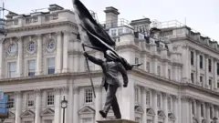 Uma estátua cinza de um homem vestindo um terno, caminhando para frente, carregando uma bandeira que cobre seu rosto