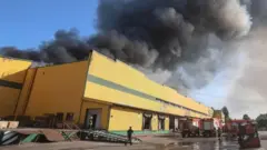 A thick, black cloud of smoke hovers above a yellow building. Fire trucks are parked in front