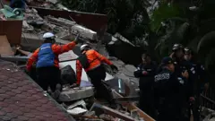 Dos rescatistas están sobre el techo de una casa colapsada, con varios policías detrás de ellos