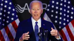 Joe Biden wears a navy suit in front of the US Flag 