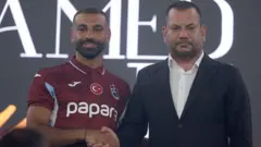 Mohamad Salah ve Trabzonspor Başkanı Ertuğrul Doğan yan yana duruyor ve el sıkışıyorlar. Salah'ın üzerinde Trabzonspor forması var. Doğan ise siyah takım elbise ve beyaz gömlek giyiyor