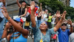 Un conjunto de personas en la calle levantan las manos y parecen saludar y estar grabando con sus teléfonos celulares. Al centro de la imagen hay una mujer con camisa azul que lleva colgada la bandera de Venezuela.