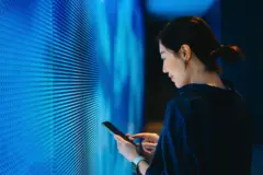 Inteligência artificial: por que robôs não conseguem resolver charadas? 3 Mulher mexe em celular em frente a parede luminosa