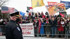 Полицейский в маске стоит перед заграждением, за которым стоит толпа людей с символикой в поддержку Дональда Трампа.